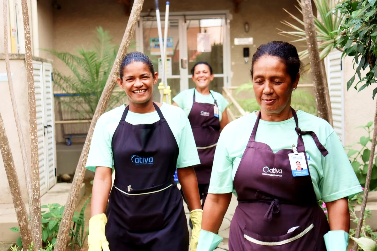 Três auxiliares de limpeza da Ativa, de avental e luvas, sorrindo na entrada de um condomínio